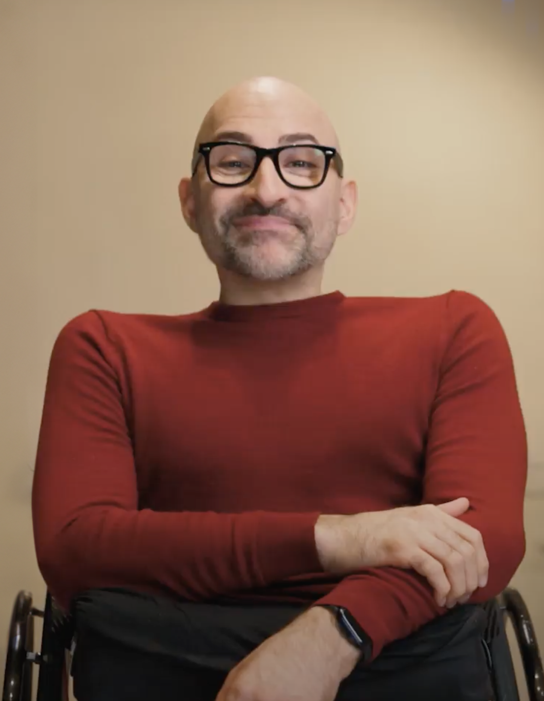 portrait of spencer west. he is a bald man wearing chunky black framed glasses, and a red sweater. he is smiling sitting in his wheelchair