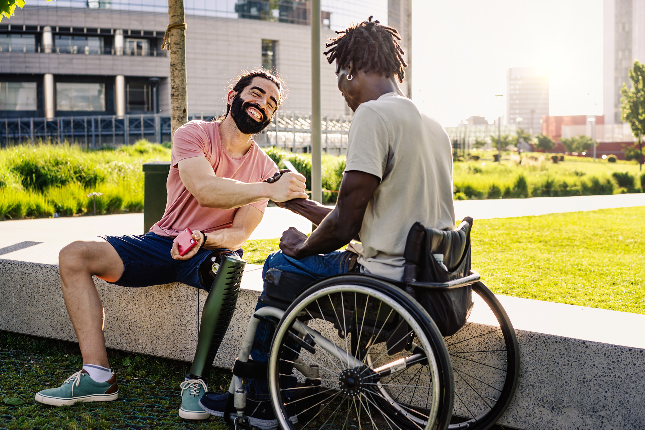 an above the leg amputee smiles and shakes hands with a wheelchair user. they are both young and attractive and clearly friends.