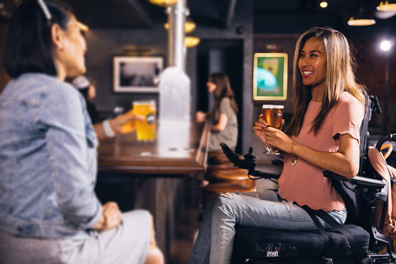 woman in power wheelchair drinks a beer with a friend at a bar