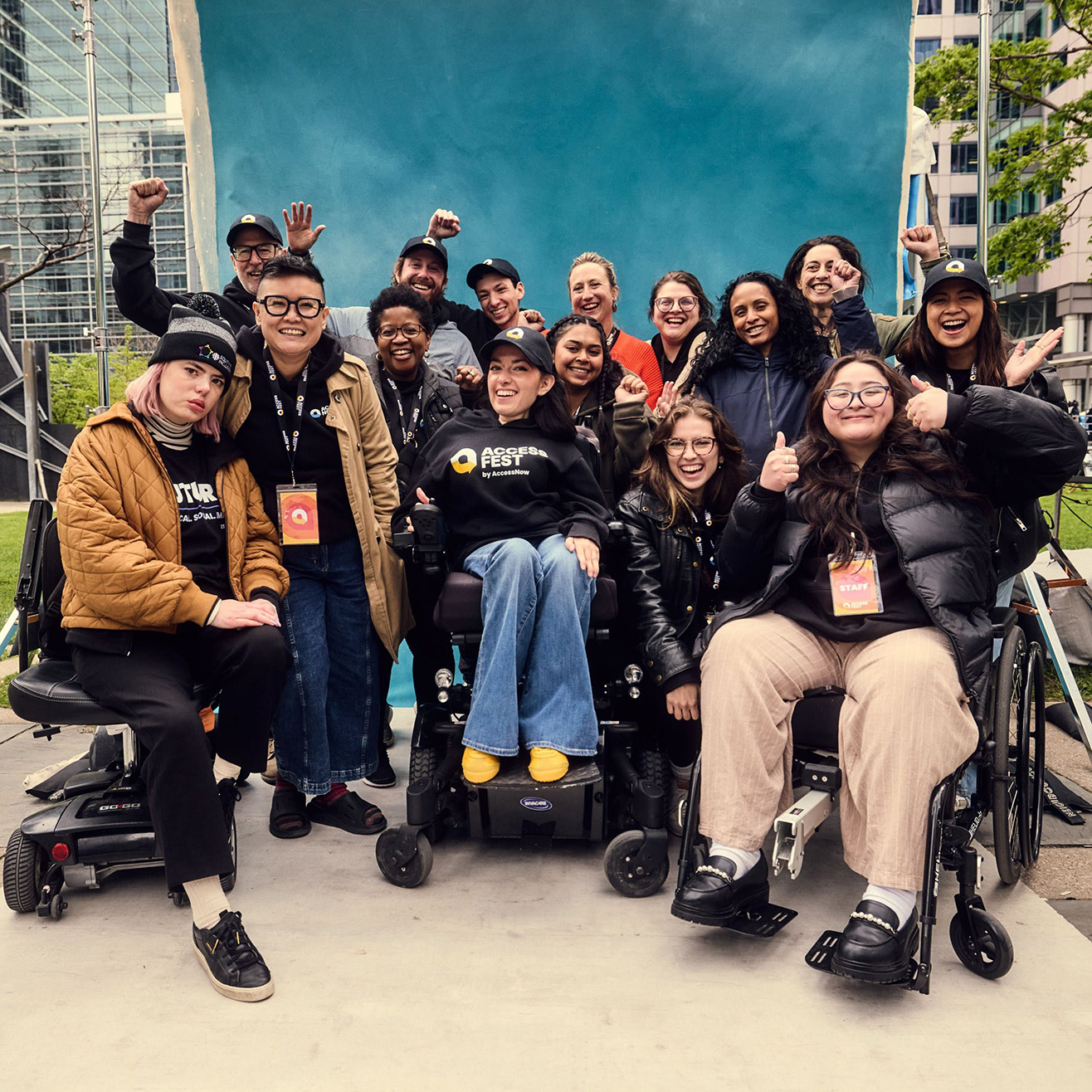 ACCESSFEST2025_093_71484 Group photo of the AccessNow team, including disabled and non-disabled individuals of diverse backgrounds, smiling and celebrating together in front of a bright blue backdrop outdoors. Several team members use wheelchairs or mobility devices
