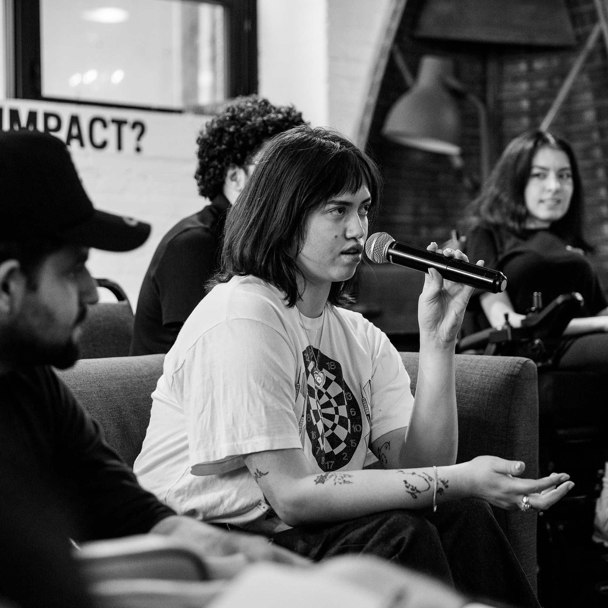 A young person with shoulder-length hair holds a microphone and speaks while seated on a couch in a cozy indoor setting. They wear a white graphic t-shirt and have visible tattoos on their arms. Other participants, including one in a power wheelchair, sit in the background, attentively listening. A partially visible poster behind them reads “IMPACT?”