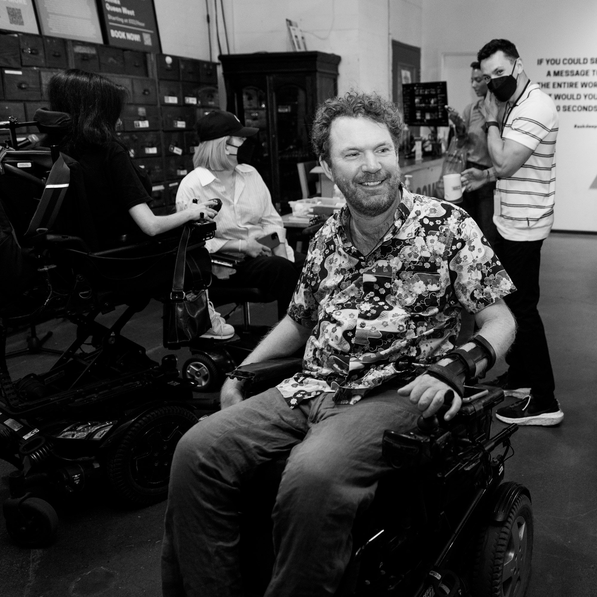 A group of people gather in a casual, creative space. A man in a patterned shirt sits in a power wheelchair, smiling and looking off to the side. Others are chatting in the background, including two people also using power wheelchairs. One person stands to the right wearing a mask and holding a drink. A large prompt on the wall asks: “If you could share a message with the entire world in 30 seconds, what would you say?”
