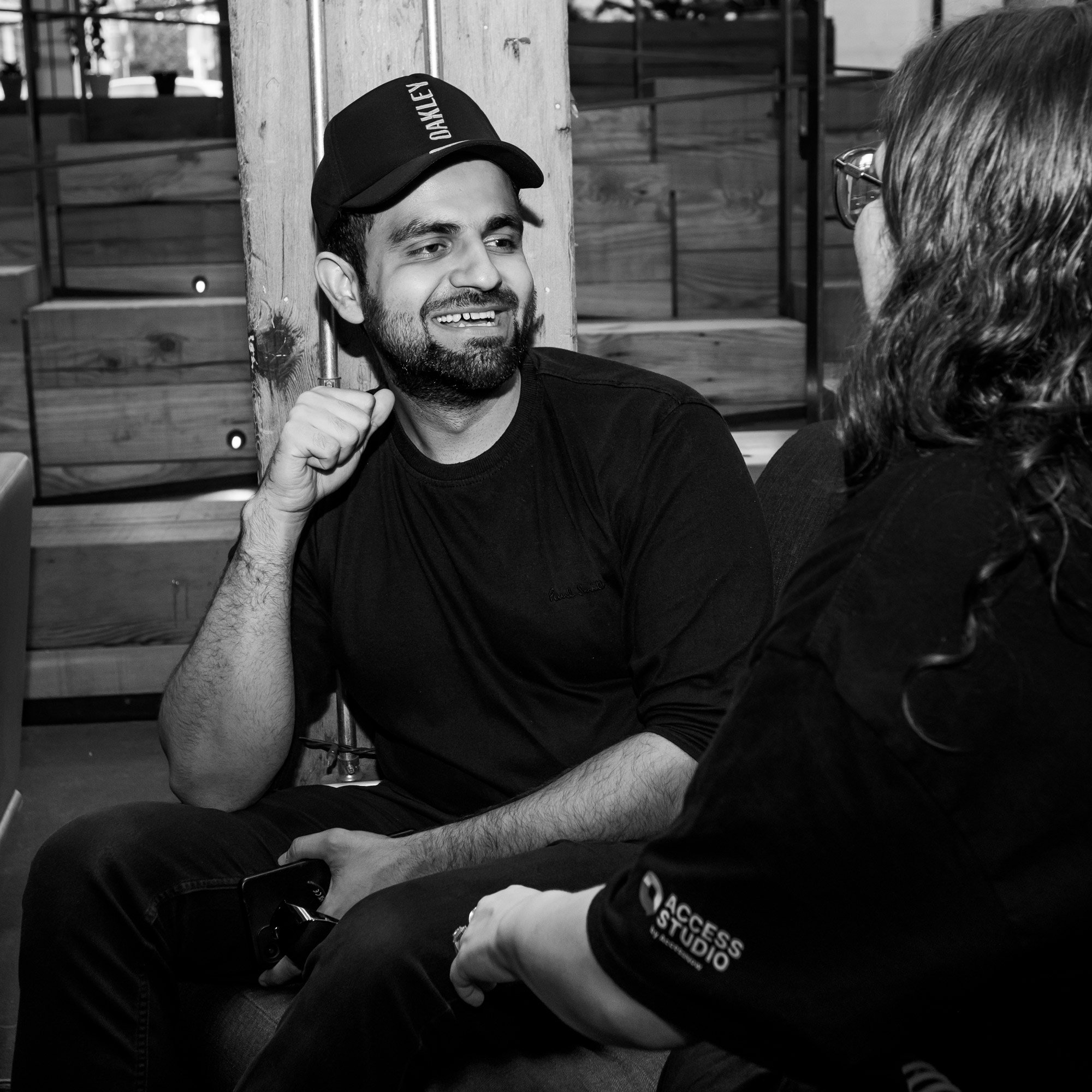 A man wearing a cap and a dark long-sleeve shirt smiles as he chats with someone out of frame. He is holding a camera and sitting comfortably in a casual lounge setting. The other person wears a shirt with the “Access Studio” logo on the sleeve.