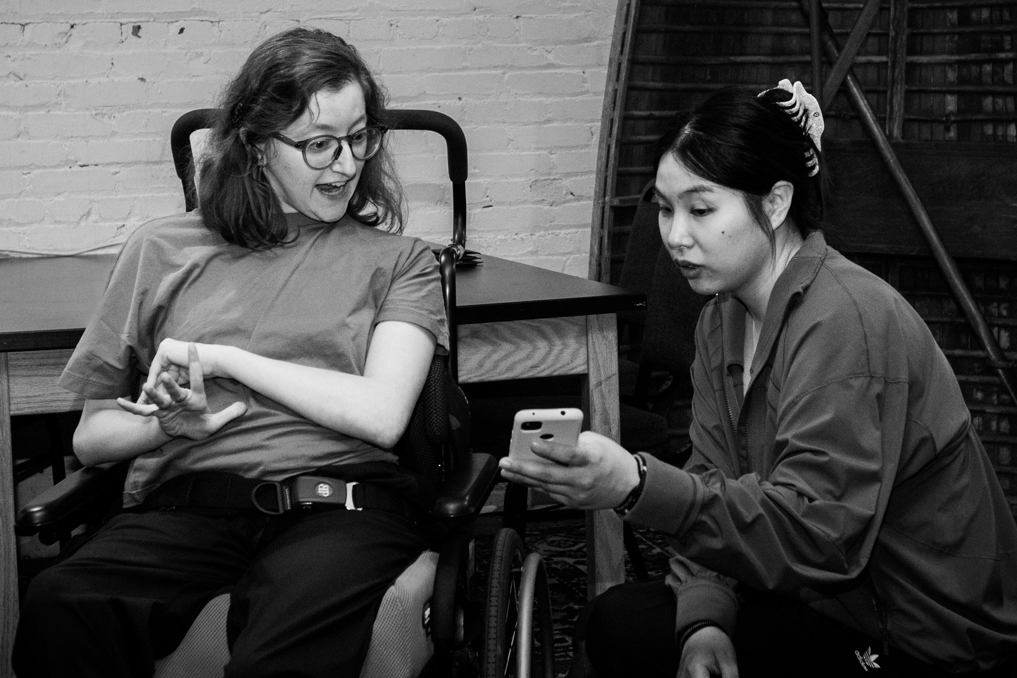 Two young people are seated indoors, closely engaged in conversation. One person, using a wheelchair and wearing glasses, appears animated and expressive, gesturing with their hands. The other, seated beside them and holding a smartphone, attentively shows them something on the screen.