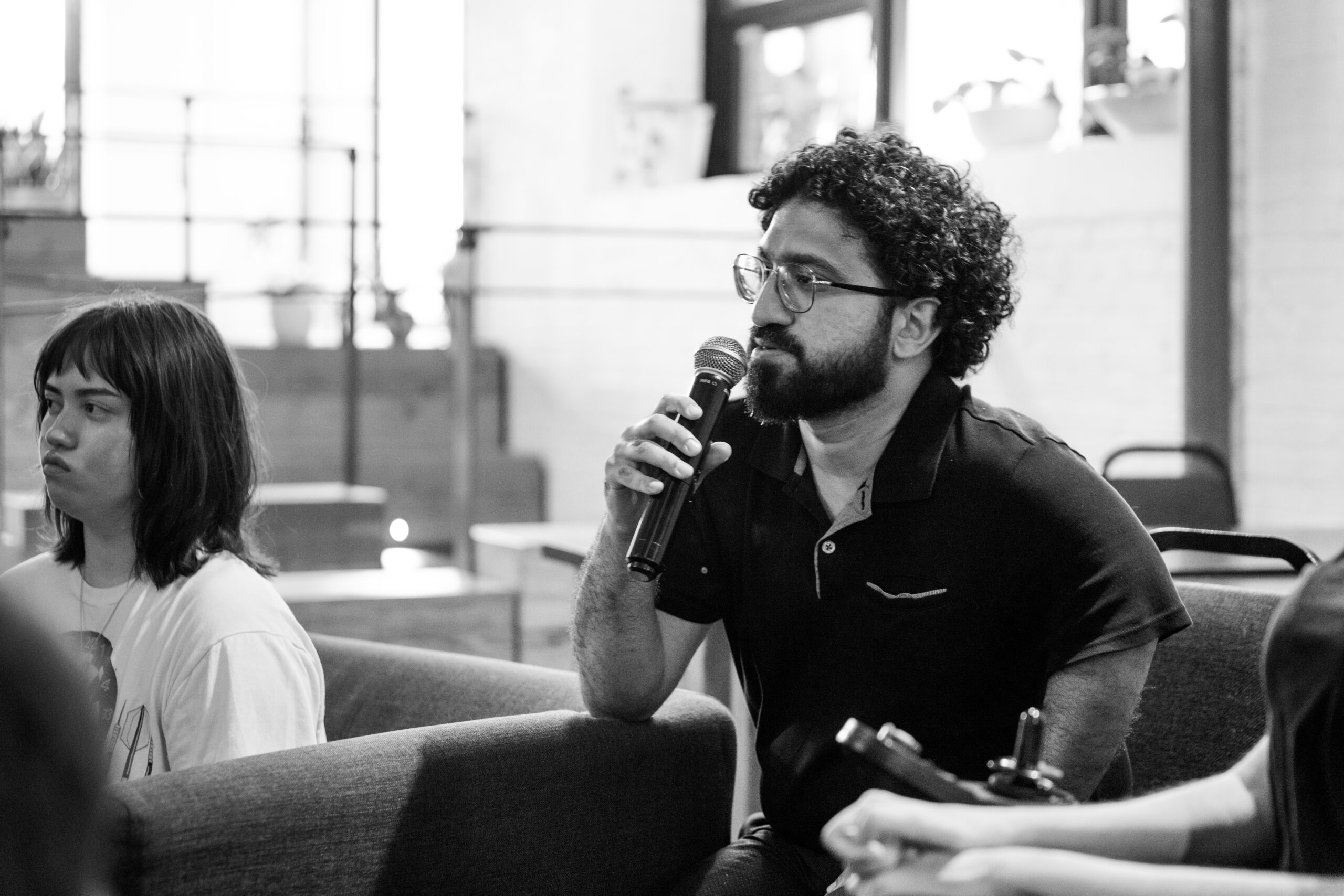Man with curly hair and glasses speaks into a microphone during a group discussion, seated beside others in a casual indoor setting.