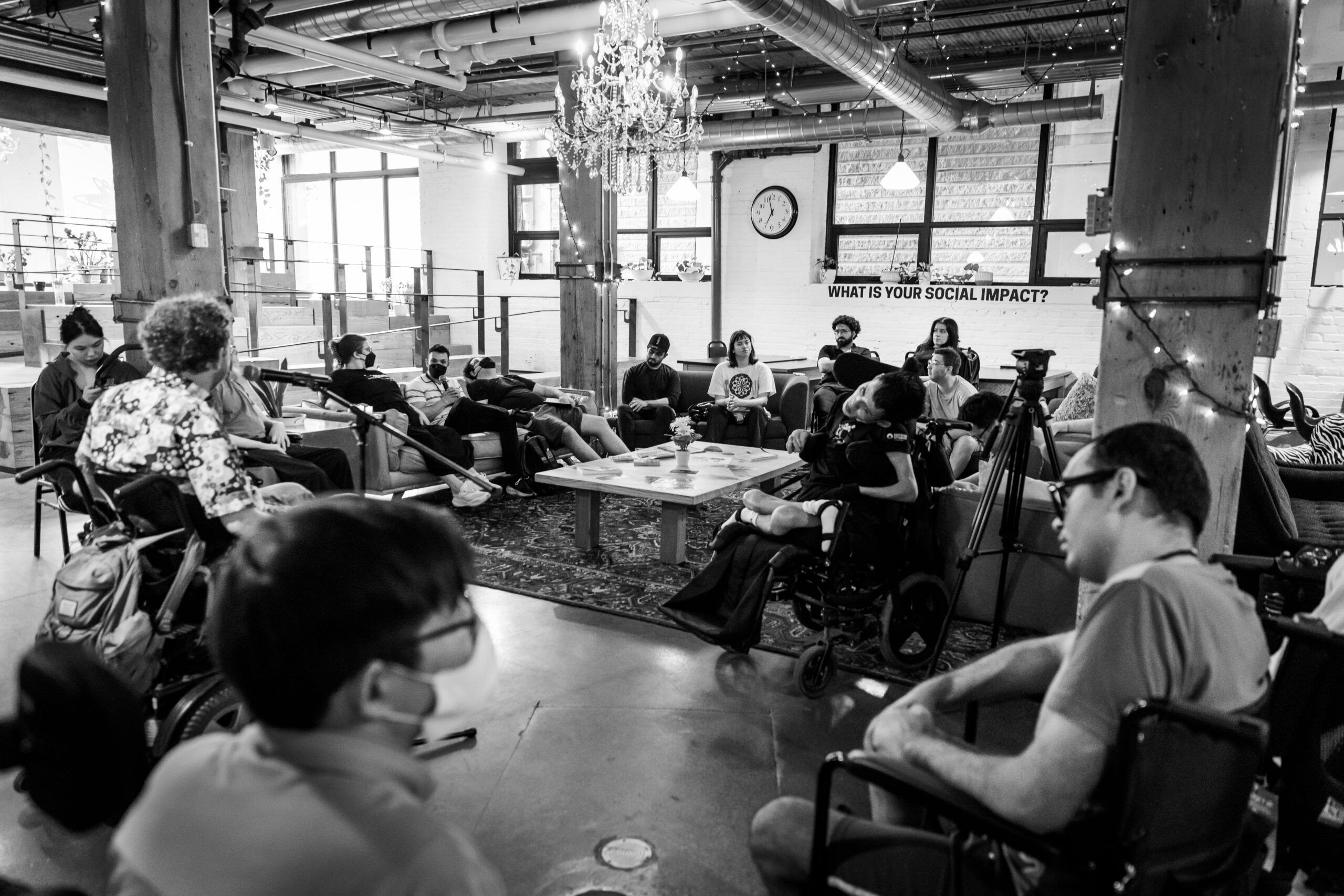 Group discussion in a loft-style space with people seated in a circle, including several wheelchair users, gathered around a speaker with a microphone; a sign on the wall reads “What is your social impact?”