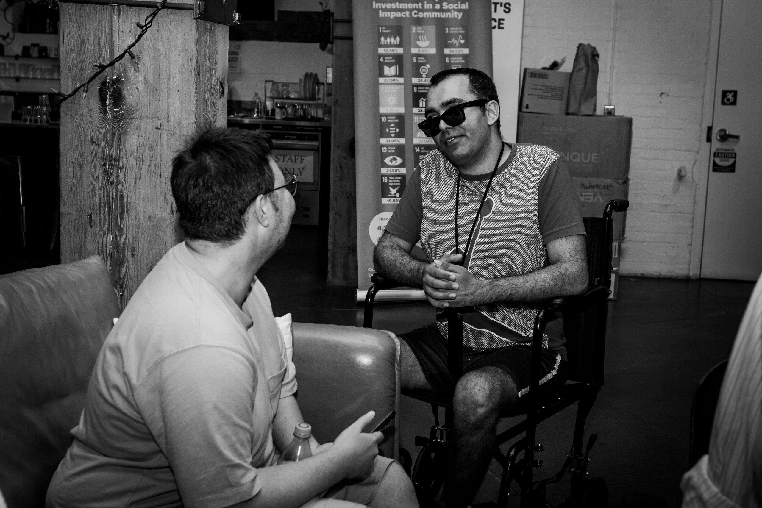 Two people in conversation in a casual indoor space, one seated in a wheelchair wearing sunglasses, as they talk face-to-face.