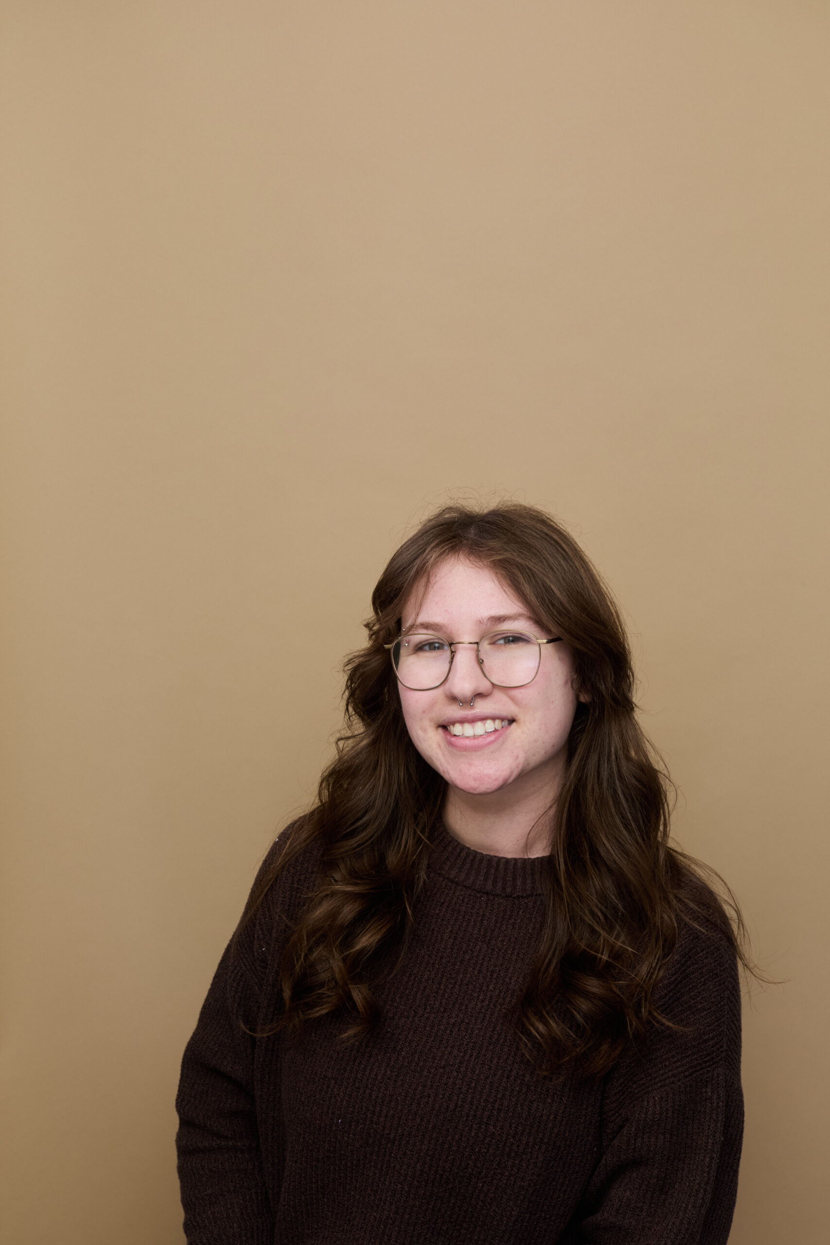 Bethany Schaufler-Biback: a smiling woman with long wavy brown hair, wearing round wire-rimmed glasses and a cozy brown knitted sweater.