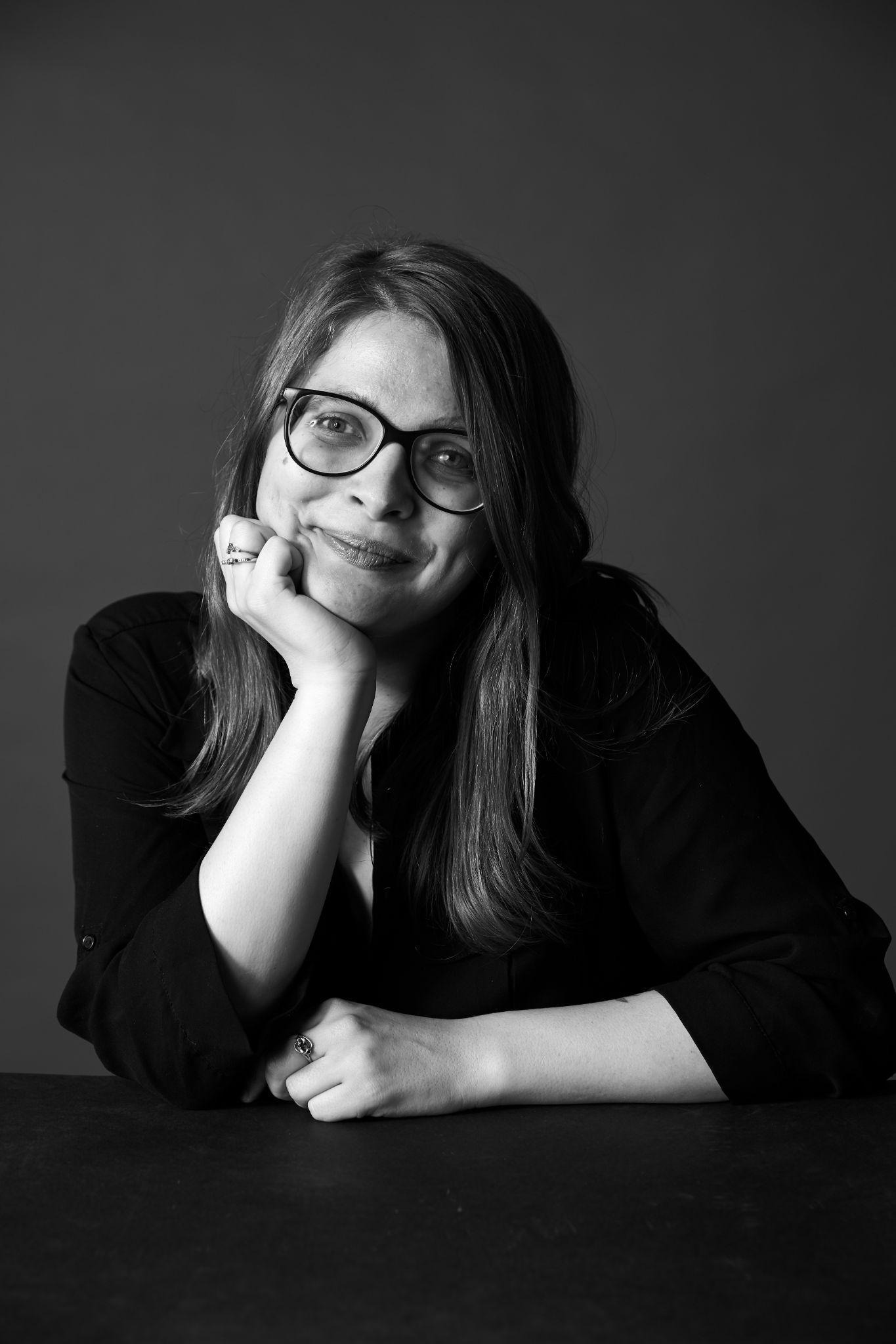 Black and white photograph of Jess, a white woman with glasses and long brown hair resting her head on her hand where she wears rings. She smiles at the camera