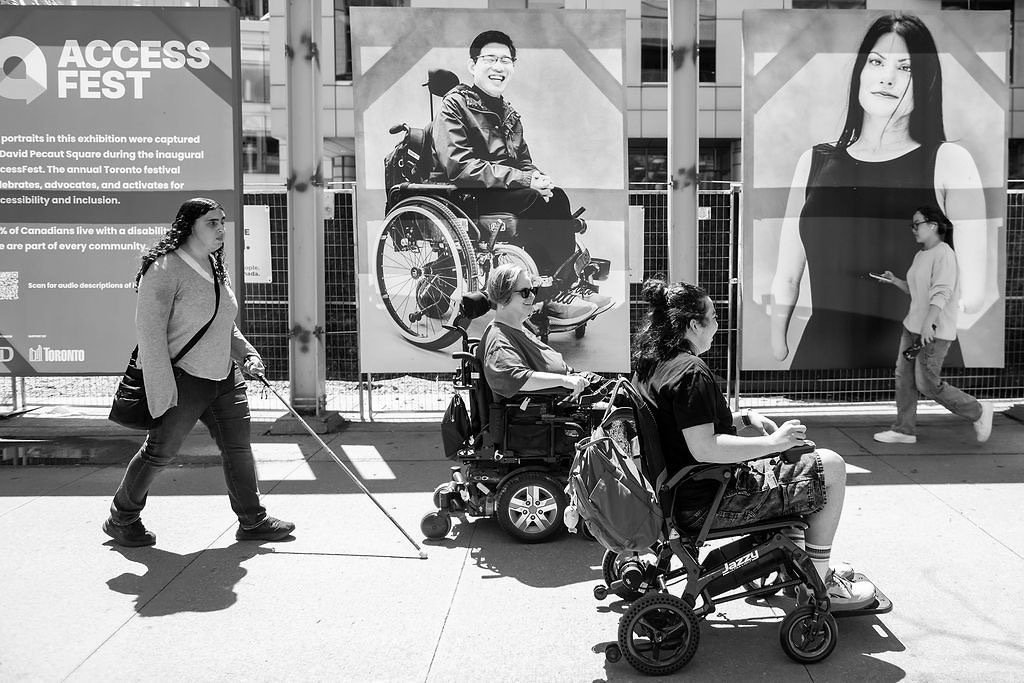 Three people move through the AccessFest portrait exhibition in Toronto. A woman using a white cane walks on the left. Two wheelchair users pause in front of large portrait photographs mounted on a fence. An AccessFest banner and City of Toronto branding are visible in the background.