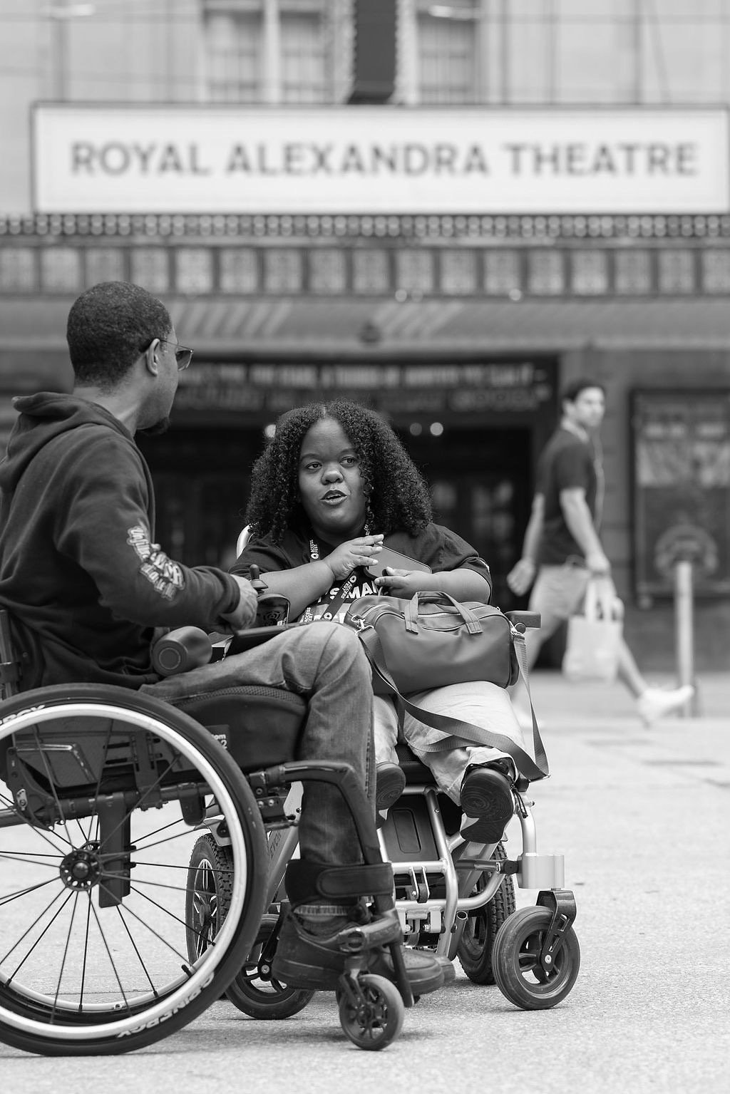 A young Black woman using a power wheelchair speaks animatedly with someone outside the Royal Alexandra Theatre in Toronto. She is wearing an AccessNow lanyard and appears mid-conversation.