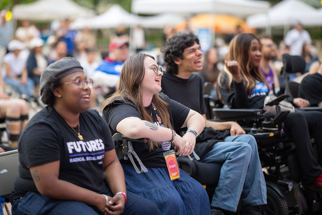 A group of FUTURES participants laugh together outdoors at an event. Several are using power wheelchairs and wearing FUTURES t-shirts. A large crowd is visible in the background.