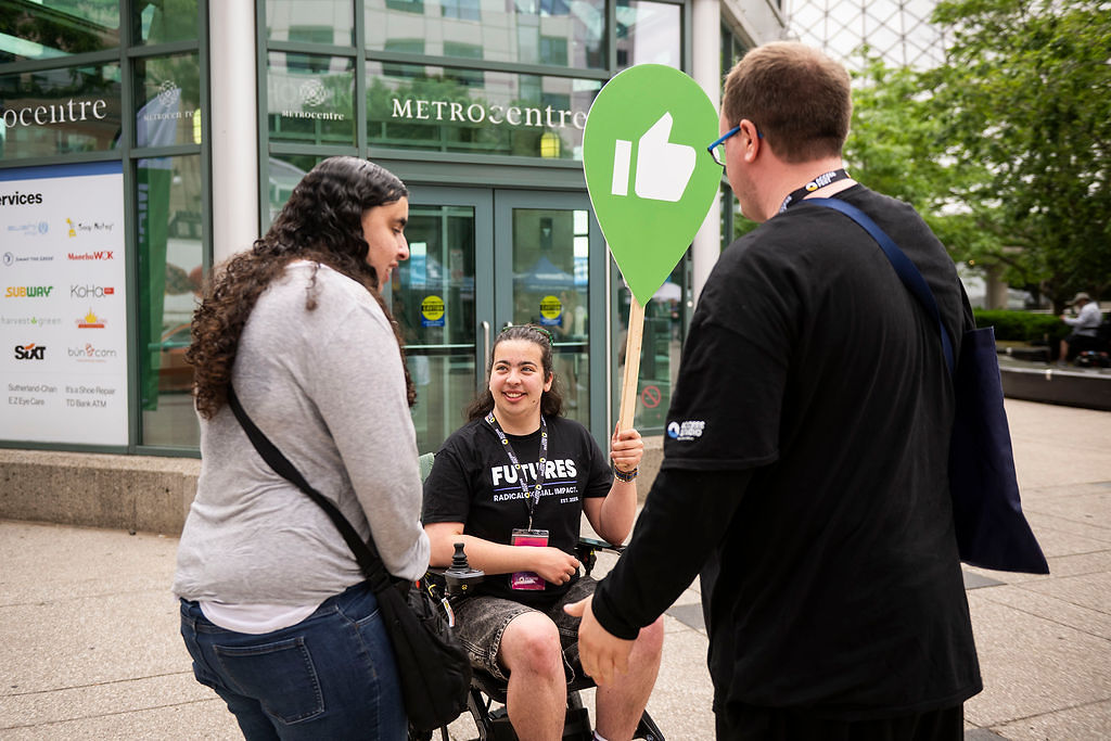 A young man in a power wheelchair, wearing a FUTURES t-shirt, smiles and holds a large thumbs-up sign while talking with two people outdoors.