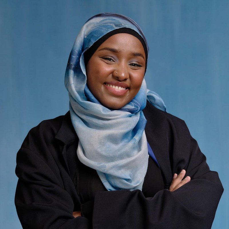 A Black woman wearing a patterned blue hijab against a blue background, smiling with her arms crossed, wearing a blazer with a QR code on the right sleeve.