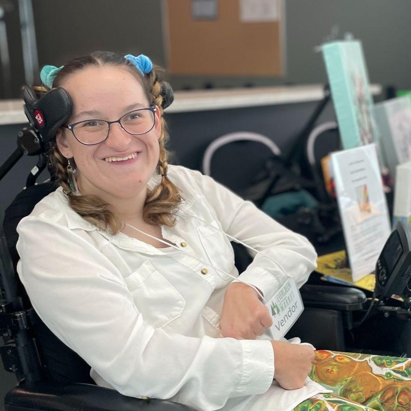 A smiling young woman with braided light brown hair, blue scrunchies, and glasses sits in a power wheelchair. She wears a white shirt and colourful pour-painted leggings beside a table of her dog paintings.